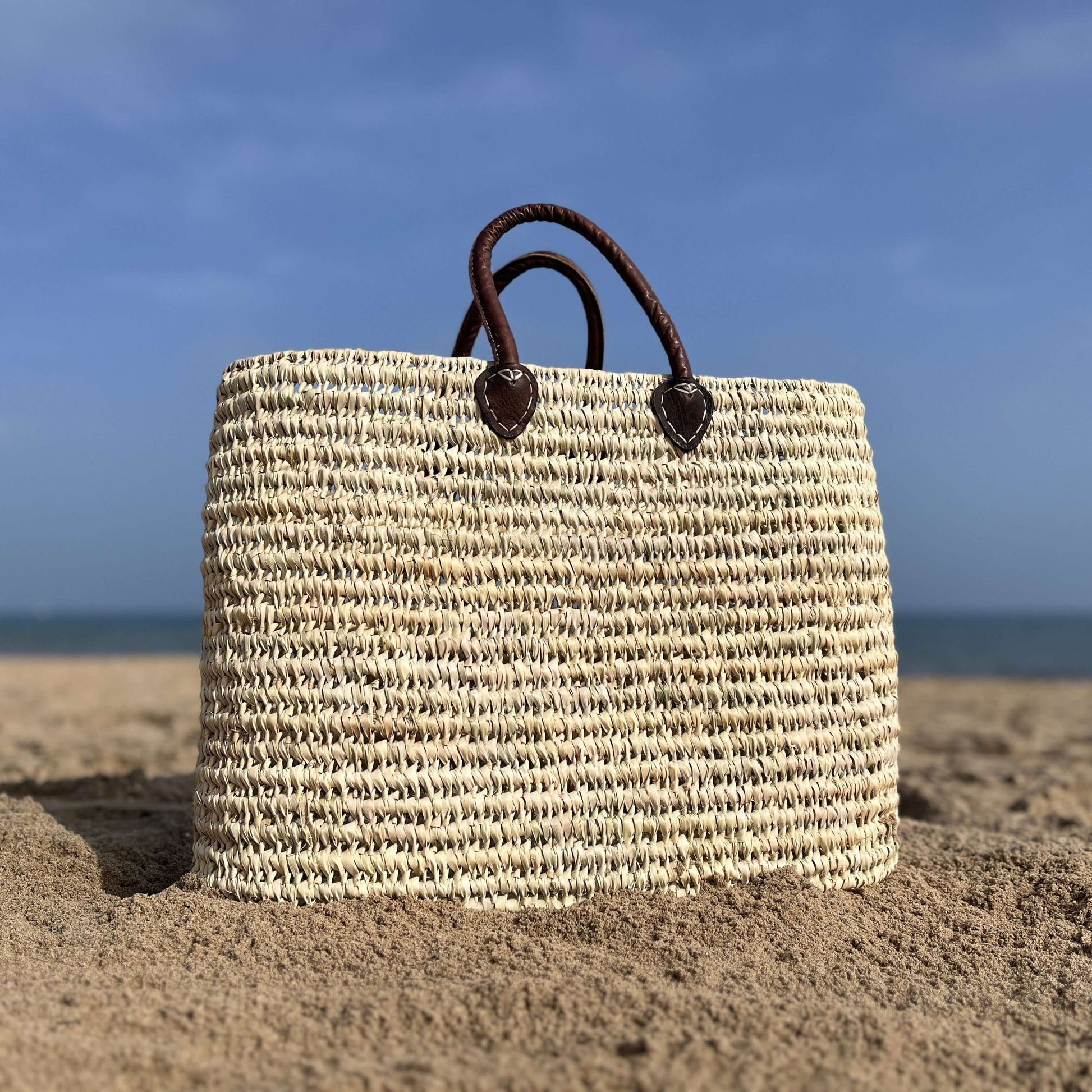 Capazo de palma grande y original con asas de cuero en la playa, ideal para almacenar artículos esenciales.